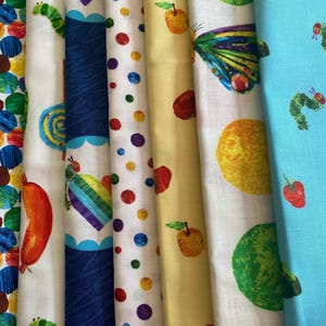Puede incluir: Una colección de rollos de tela coloridos con varios diseños. Los diseños incluyen un pájaro rojo, círculos coloridos, una oruga, manzanas y una mariposa. Las telas son principalmente blancas, azules y amarillas.