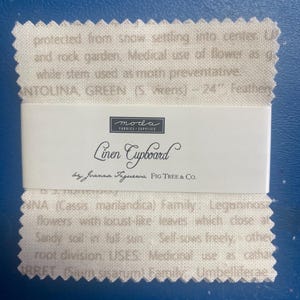 May include: A stack of fabric swatches with a white label that reads "Linen Cupboard" by Joanna Figueroa, FIG TREE & CO. The fabric is a light beige colour with text printed on it. The text includes botanical information and the words "ANTOLINA, GREEN".