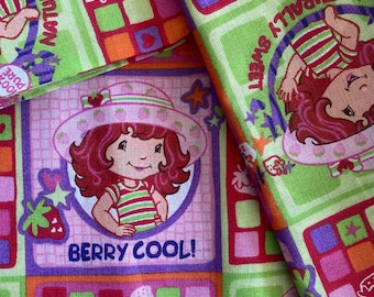 Könnte beinhalten: Nahaufnahme eines bunten Stoffes mit einem sich wiederholenden Muster einer Zeichentrickfigur mit roten Haaren und Hut, zusammen mit dem Text "BERRY COOL!". Das Design umfasst Erdbeeren, Quadrate und die Worte "NATURALLY SWEET".