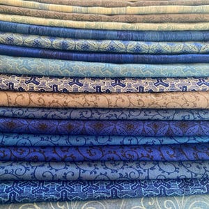 Puede incluir: Una pila de telas dobladas en varios tonos de azul y beige. Las telas muestran una variedad de patrones, incluyendo diseños florales y geométricos. La paleta de colores incluye azules claros, azul marino y beige apagado, creando una disposición visualmente atractiva.