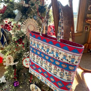 May include: A rectangular tote bag featuring a repeating pattern of Raggedy Ann and Andy dolls in blue and red. The bag has a red interior and matching handles. The bag is hanging on a wooden chair near a decorated Christmas tree.