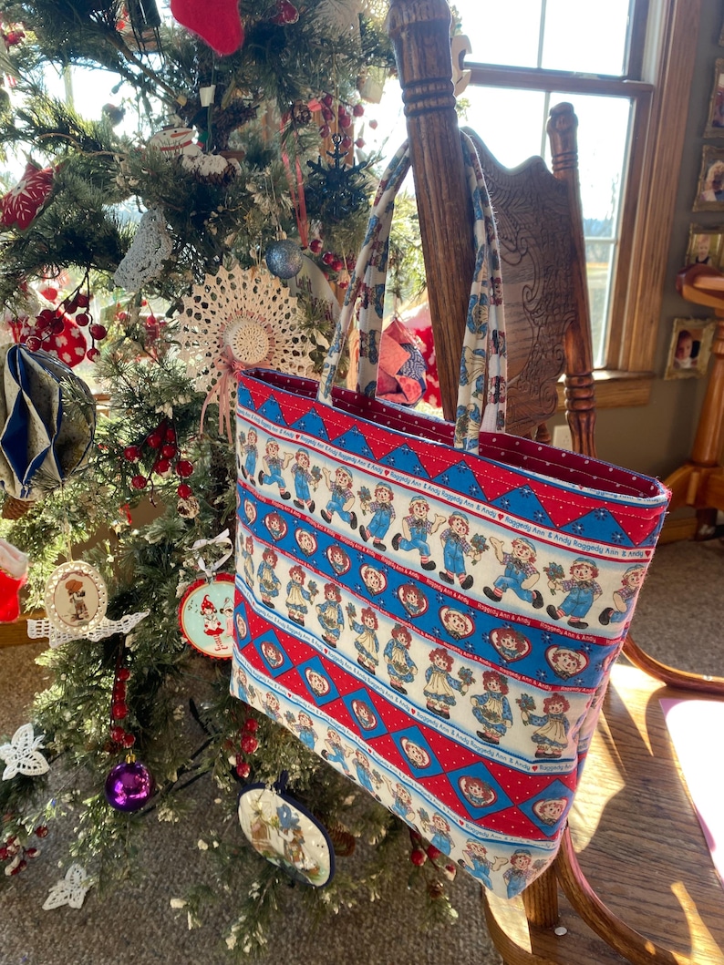 May include: A rectangular tote bag featuring a repeating pattern of Raggedy Ann and Andy dolls in blue and red. The bag has a red interior and matching handles. The bag is hanging on a wooden chair near a decorated Christmas tree.