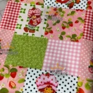 May include: A patchwork fabric featuring various patterns, including red gingham, white polka dots, and strawberry motifs. The design incorporates images of a cartoon character with a strawberry hat. The color palette includes pink, red, green, and white.