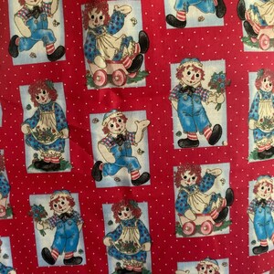 May include: Red fabric with a white polka dot pattern featuring repeating images of a Raggedy Ann doll. The doll is depicted in various poses, wearing a blue outfit, apron, and a bonnet. Some images show the doll with a bouquet or in a toy wagon.