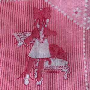 以下が含まれることがあります： ピンクと白の生地に繰り返しのパターン。パターンには、髪にリボンをつけた女の子が、猫と一緒に長椅子に座っている様子が含まれています。背景には縦縞と花の正方形があります。