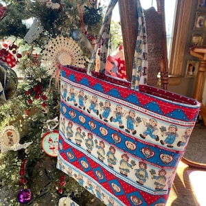 May include: A tote bag with a red interior and handles. The exterior features a repeating pattern of Raggedy Ann and Andy dolls on a white background, with red and blue accents. The bag is displayed near a decorated Christmas tree.