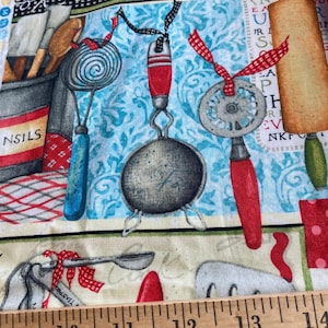 May include: A blue and white fabric with a pattern of kitchen utensils and the text "Bake with Love". The fabric is measured with a wooden ruler.