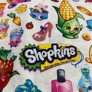 May include: A colorful fabric with a repeating pattern of Shopkins characters. The characters are cartoon-like and include a strawberry, a cupcake, a corn cob, a donut, a nail polish bottle, and a shoe. The fabric has a white background and the text "Shopkins Once you shop... You can't stop!" is printed on the fabric.