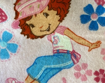 Könnte beinhalten: Stoff mit einer Zeichentrickfigur mit roten Haaren, die einen rosa Schirm, ein weißes und rosa Hemd und eine blaue Hose trägt. Der Hintergrund ist weiß mit rosa Blumen, blauen Blumen, Schmetterlingen und Erdbeeren.