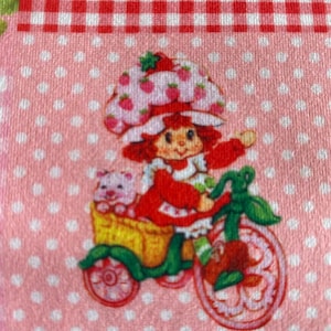 May include: A fabric featuring a Strawberry Shortcake character riding a bicycle with a pig in a basket. The design includes a red and white gingham pattern and a pink polka dot background. The character is wearing a red dress and a strawberry hat.