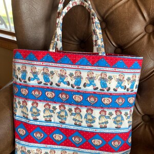 May include: A rectangular tote bag with a colorful Raggedy Ann and Andy print. The bag features a red, white, and blue pattern with images of the characters and heart shapes. The handles are patterned with a similar design.