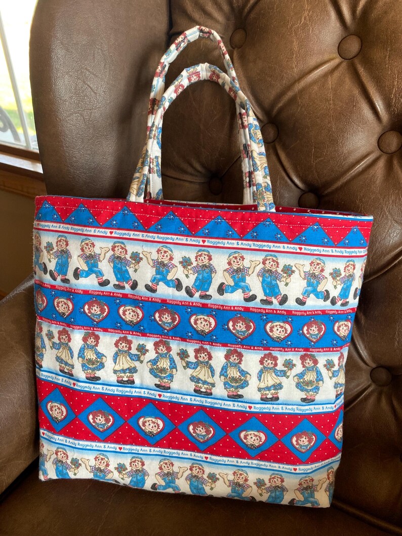 May include: A rectangular tote bag with a colorful Raggedy Ann and Andy print. The bag features a red, white, and blue pattern with images of the characters and heart shapes. The handles are patterned with a similar design.