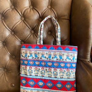 May include: A rectangular tote bag with a repeating pattern of cartoon characters in blue and red. The bag has a red and blue border with a white background. The handles are patterned with a similar design. The bag is sitting on a brown leather chair.