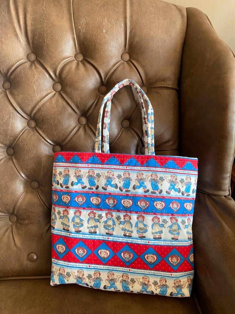 May include: A rectangular tote bag with a repeating pattern of cartoon characters in blue and red. The bag has a red and blue border with a white background. The handles are patterned with a similar design. The bag is sitting on a brown leather chair.