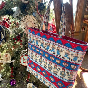 May include: A rectangular tote bag with a repeating pattern of Raggedy Ann and Andy dolls on a white background, framed by red and blue geometric designs. The bag has matching patterned handles and a red interior. The bag is hanging near a decorated Christmas tree.