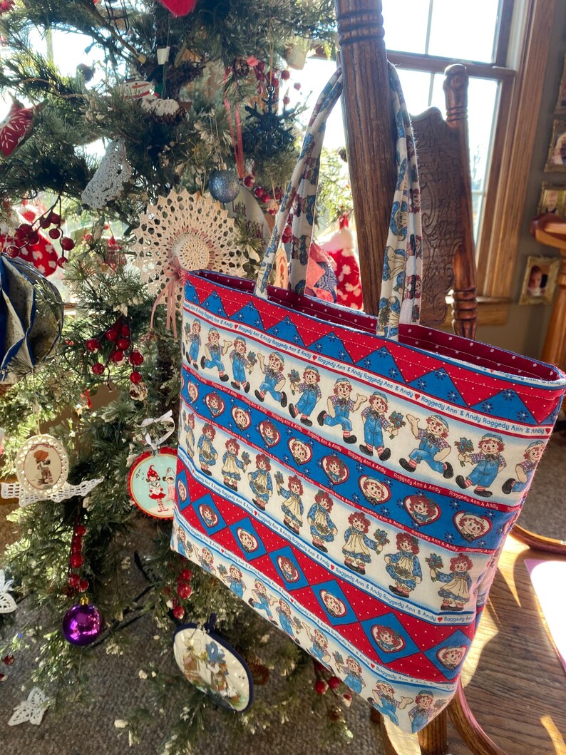 May include: A rectangular tote bag with a repeating pattern of Raggedy Ann and Andy dolls on a white background, framed by red and blue geometric designs. The bag has matching patterned handles and a red interior. The bag is hanging near a decorated Christmas tree.