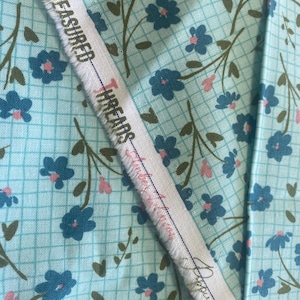 以下が含まれることがあります： 水色の背景に格子模様のフローラルファブリックのクローズアップ。生地は、ピンクの中心と茶色の茎を持つ青い花を特徴としています。「TREASURED THREADS」というテキストが入った白いストリップが見えます。