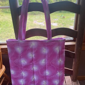 Puede incluir: Un bolso tote rosa con un estampado de hadas. El bolso tiene asas largas y está hecho de tela acolchada. El diseño de hadas presenta siluetas blancas y líneas punteadas sobre un fondo rosa.