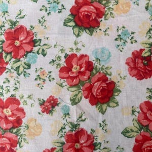 May include: Close-up of a white fabric with a repeating floral pattern. Large red roses are interspersed with smaller blue and yellow flowers and green leaves. The fabric has a vintage, cottagecore aesthetic.
