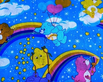 May include: A blue fabric featuring a repeating pattern of Care Bears characters, including yellow, purple, pink, and blue bears. The design includes rainbows, white clouds, pink and brown heart-shaped balloons, and yellow stars scattered across the background.