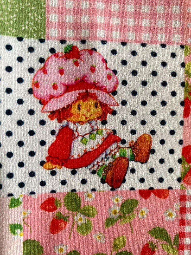 May include: A patchwork design featuring a Strawberry Shortcake character on a white polka dot background. The character wears a pink strawberry hat and red outfit. The patchwork includes pink gingham, green floral, and strawberry patterns.