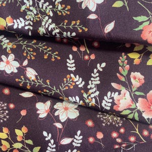 Puede incluir: Primer plano de una tela con un estampado floral. La tela es de color púrpura oscuro y presenta un diseño repetido de flores y follaje blancos, melocotón y amarillos. La tela está doblada, mostrando la textura y la profundidad del material.