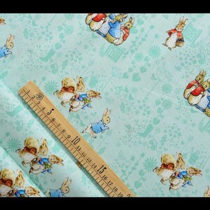 May include: A light blue fabric with a repeating pattern of Peter Rabbit characters. The fabric is measured with a ruler that shows 19 inches.