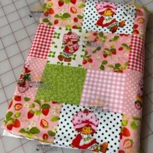 May include: A patchwork fabric item featuring various patterns, including strawberries, gingham, polka dots, and a cartoon character. The color palette includes pink, red, green, and white. The item appears to be a blanket or quilt.
