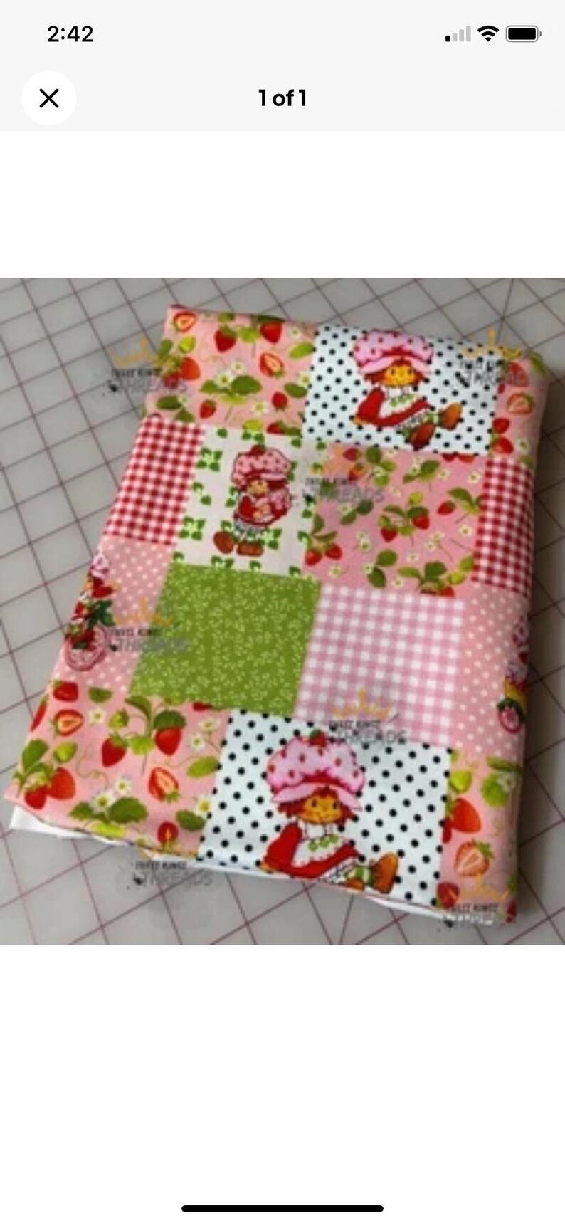May include: A patchwork fabric item featuring various patterns, including strawberries, gingham, polka dots, and a cartoon character. The color palette includes pink, red, green, and white. The item appears to be a blanket or quilt.