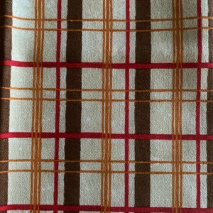May include: Close-up of a fabric with a plaid pattern. The design features a beige background with intersecting lines in brown, red, and orange. The fabric has a textured appearance, suggesting a woven material.
