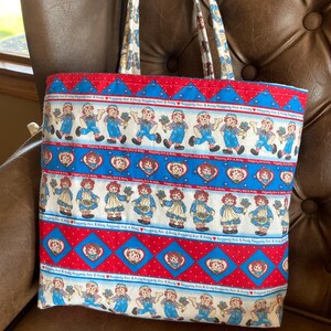 May include: A fabric tote bag featuring a repeating pattern of Raggedy Ann and Andy dolls in blue overalls and dresses, set against a red and blue background. The bag has matching handles and a sturdy construction, perfect for carrying items.