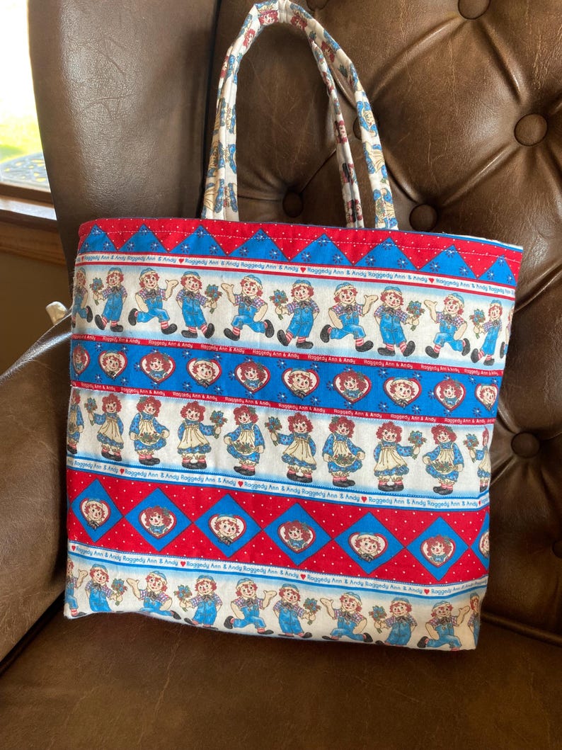 May include: A fabric tote bag featuring a repeating pattern of Raggedy Ann and Andy dolls in blue overalls and dresses, set against a red and blue background. The bag has matching handles and a sturdy construction, perfect for carrying items.