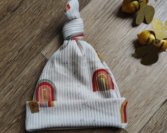 May include: A white baby hat with a knotted top and a rainbow pattern. The hat has a small brown label with the text "Boho" on it.