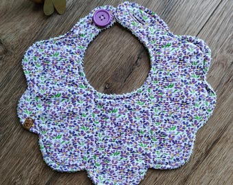 Könnte beinhalten: Ein Baby-Lätzchen mit lila und weißem Blumenmuster, verschlossen mit einem lila Knopf. Das Lätzchen hat einen gewellten Rand und ist aus weichem Stoff gefertigt. Ideal für Babys beim Essen.