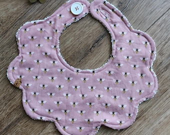 Könnte beinhalten: Rosa Baby-Lätzchen mit einem sich wiederholenden Muster aus schwarzen und gelben Bienen. Das Lätzchen hat einen gewellten Rand und einen weißen Knopfverschluss. Das Lätzchen ist aus Stoff und soll Essensspritzer auffangen.