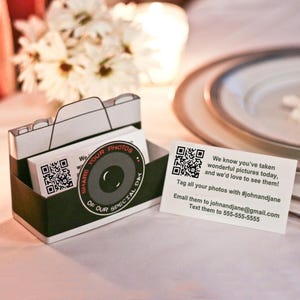 May include: A camera-shaped paper card holder in black and white with a circular lens graphic that reads SHARE YOUR PHOTOS OF OUR SPECIAL DAY. Beside it, a white rectangular card features a QR code and text: We know you've taken wonderful pictures today, and we'd love to see them! Tag all your photos with #johnandjane. Email them to johnandjane@gmail.com. Text them to 555-555-5555.