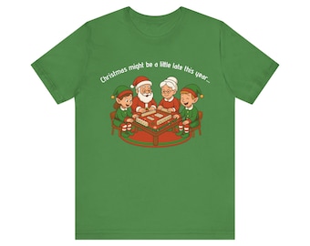 Könnte beinhalten: Grünes T-Shirt mit einer Cartoon-Illustration von Santa, Mrs. Claus und zwei Elfen, die ein Spiel spielen. Der Text "Christmas might be a little late this year..." ist über den Figuren aufgedruckt.