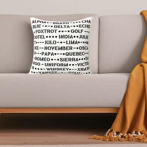 May include: A white decorative pillow with black text featuring the NATO phonetic alphabet. The pillow is rectangular and rests on a light-colored sofa. A mustard-colored throw blanket is draped over the sofa arm.