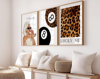 May include: Three framed wall art prints. The first print features a woman in a towel and sunglasses drinking a pink beverage, with the text "Feeling Lucky". The second print has a black 8-ball and a leopard print. The third print has a leopard print and the text "LUCKY ME".