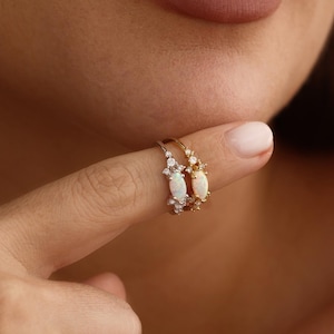 Peut inclure: Deux bagues avec une opale centrale taille marquise entourée de petites pierres claires. Une bague est en métal argenté et l'autre en métal doré. Le texte FionaGemoStudio For You est visible sur l'image.