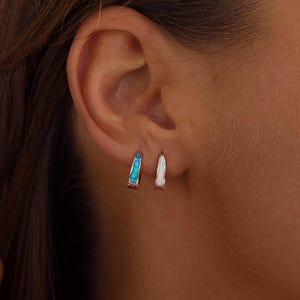 Puede incluir: Primer plano de dos pequeños pendientes de aro. Un pendiente presenta una incrustación turquesa vibrante, mientras que el otro tiene una incrustación blanca. Los pendientes se muestran contra la oreja de una persona, destacando su delicado diseño.