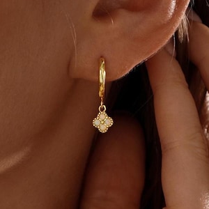 Pode incluir: Um brinco de argola dourado com um pequeno pingente de flor de quatro pétalas. O pingente é adornado com quatro pequenas pedras redondas brancas iridescentes dispostas em forma de trevo, suspenso por uma argola de metal dourado polido.