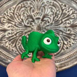 Peut inclure: Figurine de caméléon vert vif avec de grands yeux expressifs. La figurine est tenue dans une main devant un fond d'ornement mural argenté et un mur bleu. Le caméléon est un personnage de dessin animé.