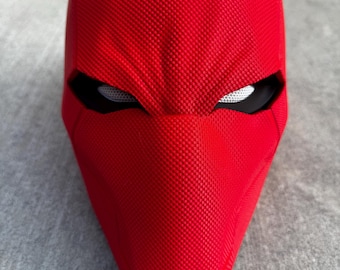 May include: A bright red, textured mask with black eye openings and white mesh eye coverings. The mask has a pointed chin and a smooth, curved top. The mask is likely a costume or cosplay item.