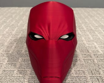 May include: A vibrant red Red Hood mask with a textured surface. The mask features a distinctive design with eye openings and a defined facial structure, perfect for cosplay or display.