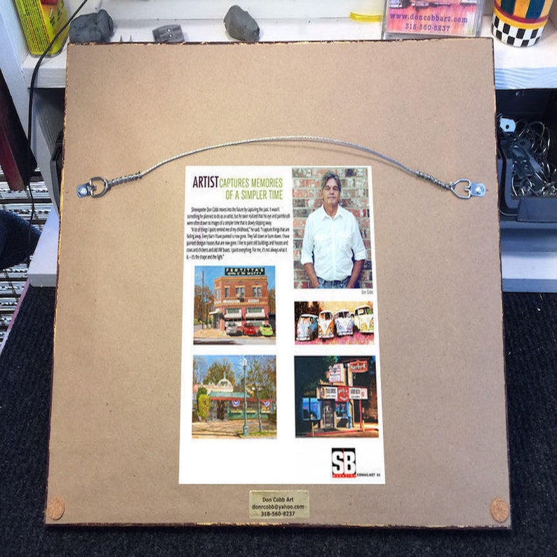 May include: The back of a framed artwork with a wire for hanging. A printed article and images are attached, featuring the artist and his work. The text includes "ARTIST CAPTURES MEMORIES OF A SIMPLER TIME" and contact information.