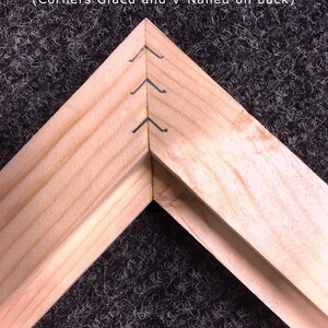May include: Close-up of a wooden frame corner, showing the construction details. The corners are glued and V-nailed for added strength. The wood has a natural, light color, and the frame is set against a dark, textured background.