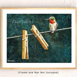 May include: A framed art print featuring a hummingbird perched on a clothesline between two wooden clothespins. The bird has green, red, and white feathers. The background is a dark teal color. The artist's signature and text "Don Cobb Art" are visible.
