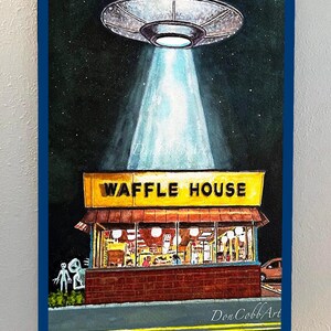 Puede incluir: Una pintura de acuarela de un OVNI flotando sobre un restaurante Waffle House por la noche. Dos extraterrestres grises est&aacute;n de pie afuera del restaurante. La pintura est&aacute; titulada "Don Cobb Art" y es una impresi&oacute;n de 61x91 cm.