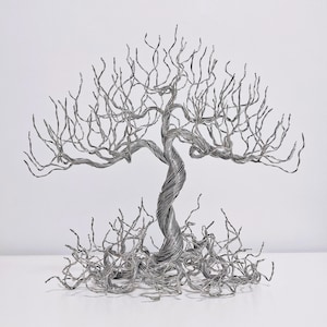 Puede incluir: Una escultura artesanal de árbol de alambre metálico. El árbol presenta un tronco retorcido y ramas intrincadas, con una copa y raíces completas. La escultura está hecha de alambre de color plateado y se encuentra sobre una superficie blanca.